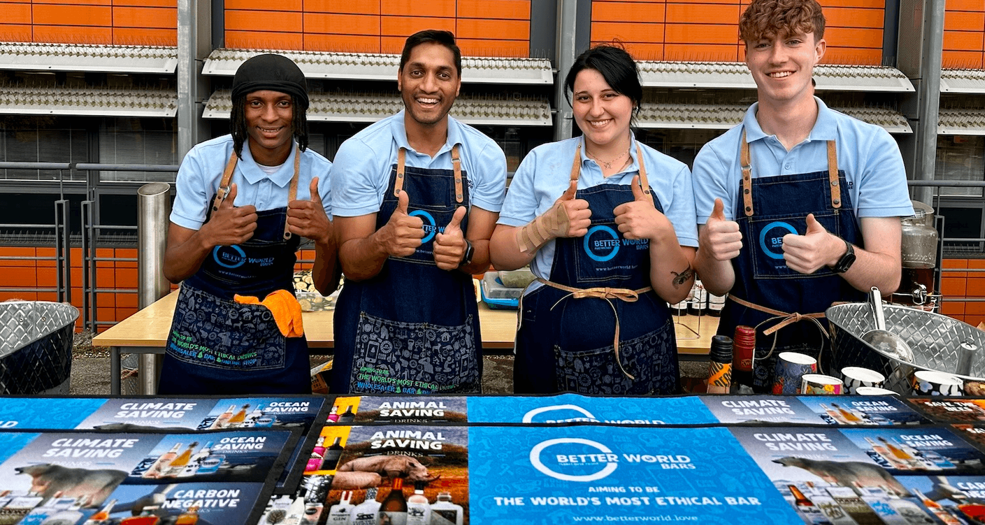 Four professional team members smiling and giving a thumbs up behind a Better World Bar Hire setup featuring climate saving branding representing our mission to redefine ethical hospitality and employ vulnerable staff