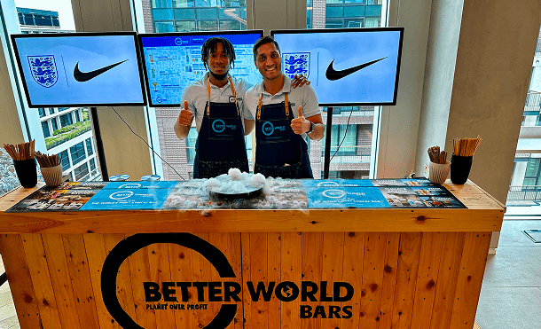 Two professional bartenders giving a thumbs up behind a sustainable wooden pop up bar featuring a dry ice display and digital screens representing our multi award winning legacy in ethical hospitality
