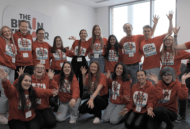 A large group of smiling charity workers wearing red The Brain Tumour Charity clothing and waving together representing our mission to inspire industry change and prove that business can build a better world