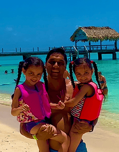 A smiling man standing in clear blue ocean water holding his two daughters who are wearing bright pink life vests with a tropical beach and pier in the background