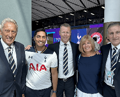 A smiling man wearing a white Tottenham Hotspur football shirt standing alongside three men in suits and a woman at a stadium representing the founder and his passion as a fan