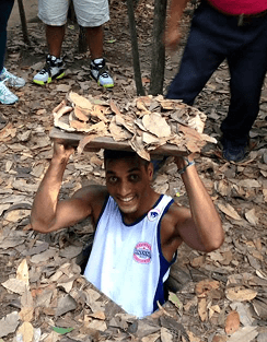 A smiling man emerging from a small camouflaged underground tunnel opening in a forest holding a leaf covered lid above his head representing his extensive world backpacking travels