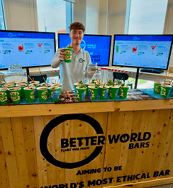 A bartender holding up a green eco disposable cup behind a wooden event bar filled with sustainable drinks