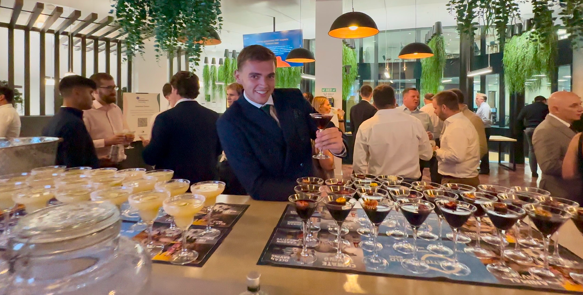 A smiling bartender in a suit holds an espresso martini behind a bar filled with prepared cocktails at a premium corporate networking event