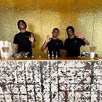 Three smiling event bartenders waving from behind a premium tiled mobile bar with a gold background