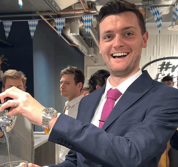 A smiling man in a suit pouring a glass of beer from a mobile event bar tap at a corporate office party