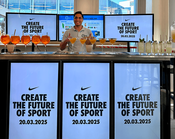 A smiling bartender giving a thumbs up behind a digital mobile event bar displaying Nike branding and lined with orange cocktails