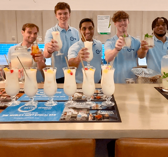 Five smiling bartenders raising their cocktail glasses in a toast behind a pop up office event bar filled with fresh drinks