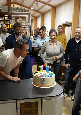 A man blowing out candles on a Certified B Corporation birthday cake while smiling guests celebrate at the Kent B Corp anniversary event featuring our premium and fully sustainable mobile bar service