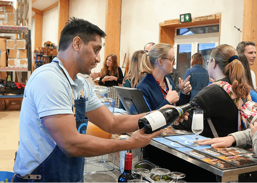 A male bartender in a branded denim apron pouring a glass of super sustainable Ridgeview English sparkling wine for guests at the B Corp certified mobile bar