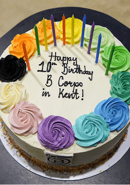 A close up view of a white birthday cake decorated with colourful icing rosettes and candles featuring the text happy 10th birthday B Corps in Kent