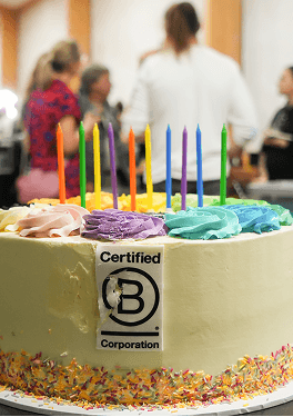 A man blowing out the colourful candles on a Certified B Corporation birthday cake surrounded by smiling guests at the Kent B Corp anniversary event