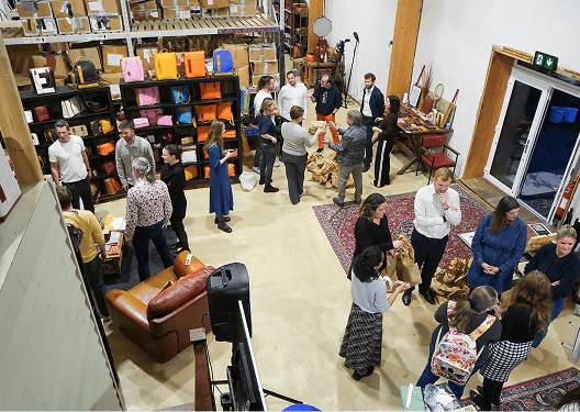 A high angle overhead view of the Kent B Corp anniversary celebration. Guests network in groups on a large area rug next to a display of upcycled products and a professional speaker setup.