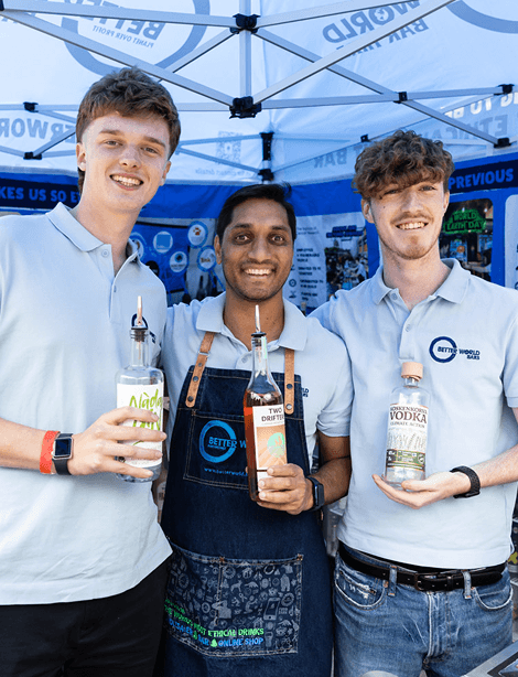 Three smiling bartenders holding sustainable spirit bottles behind a mobile event bar under a branded blue and white canopy