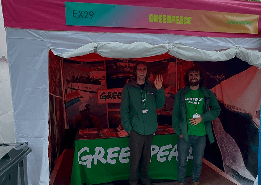 Two smiling men standing inside a Greenpeace charity tent at an outdoor sustainability festival