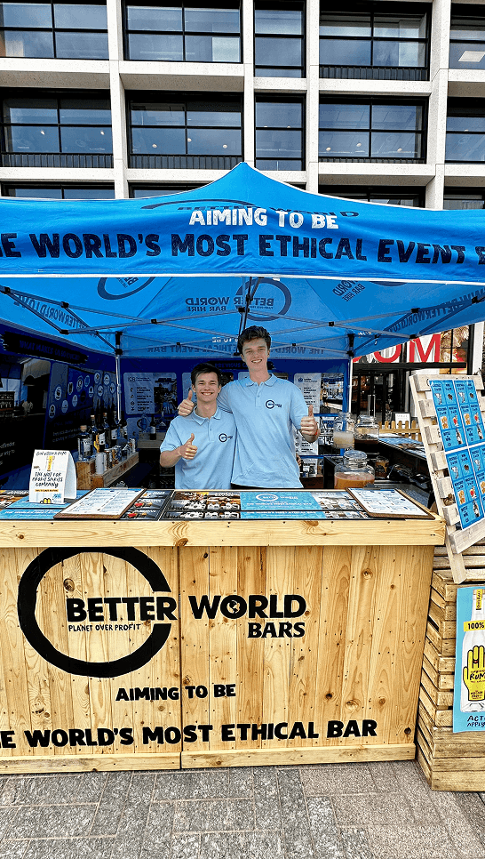 Better World Bar Hire team giving a thumbs up behind their wooden festival event bar at Earthfest
