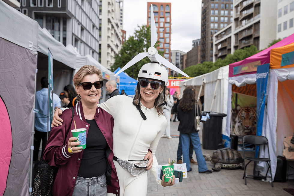 Two smiling festival attendees holding green eco friendly cups with one person wearing a white windmill hat