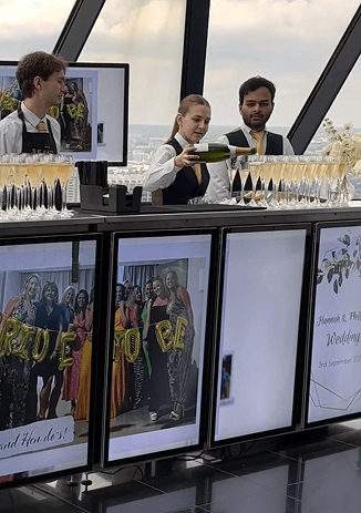Two venue staff members standing behind a long digital mobile bar displaying photos of the bride and groom against the London skyline