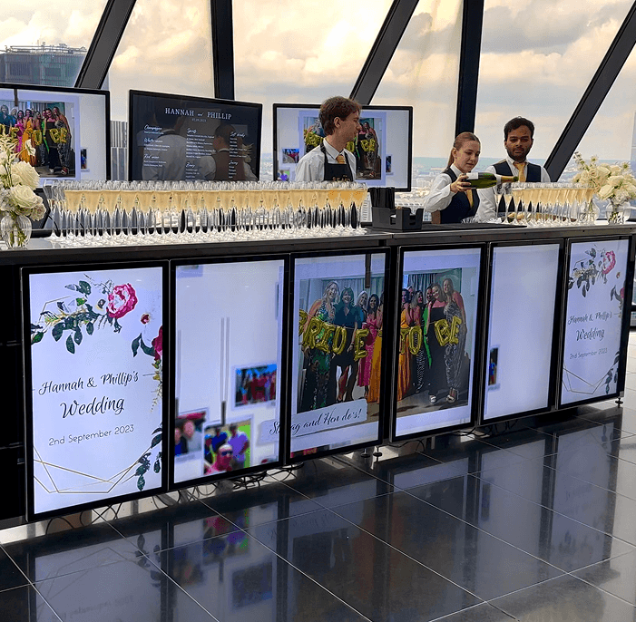 A premium digital mobile bar displaying photos of the wedding couple and a floral design setup with a long row of champagne flutes inside the Gherkin in London l l