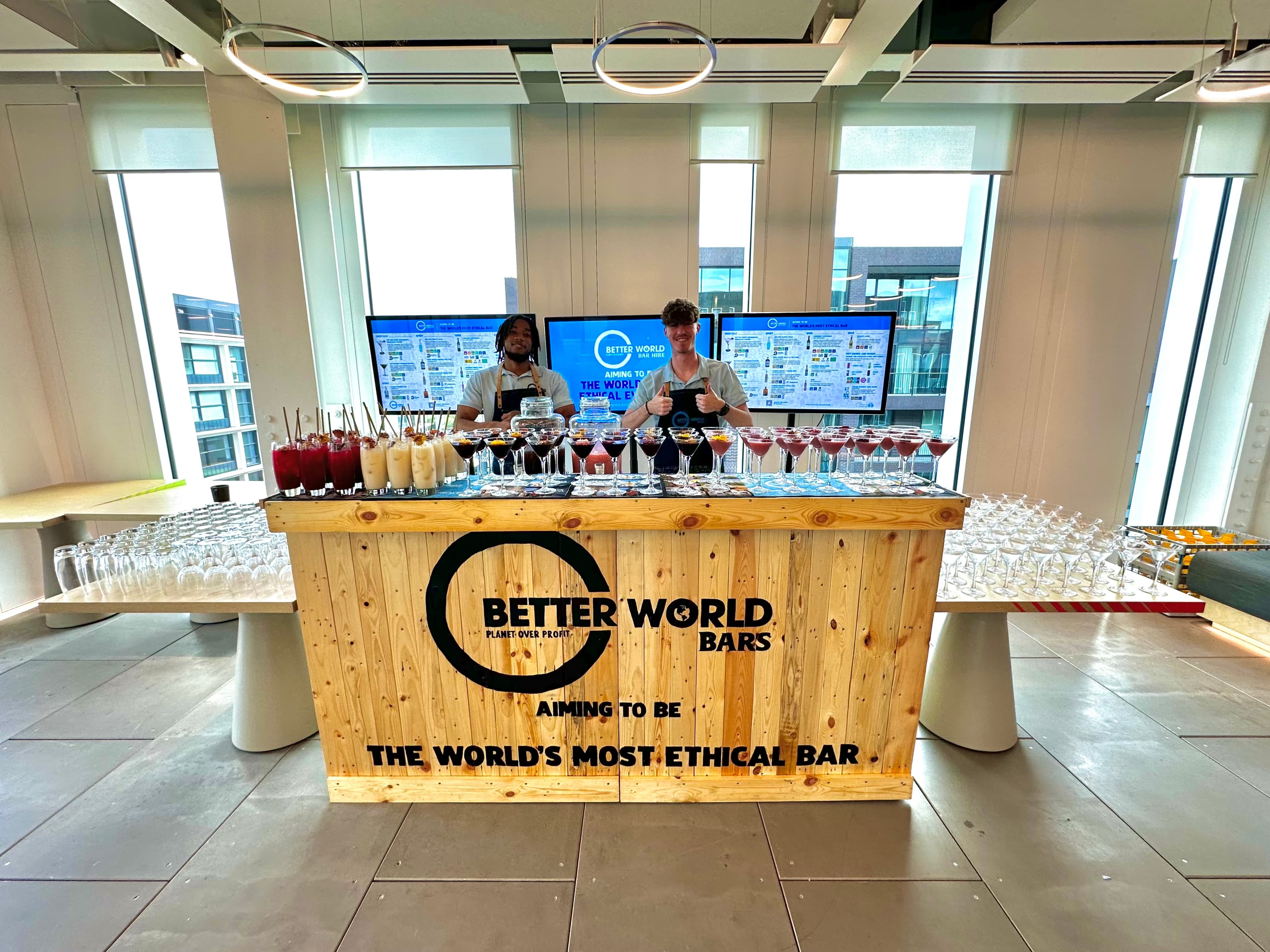 Two Better World Bar Hire bartenders behind their reclaimed wood mobile event bar, a row of cocktails poured beneath the digital drinks-menu screens
