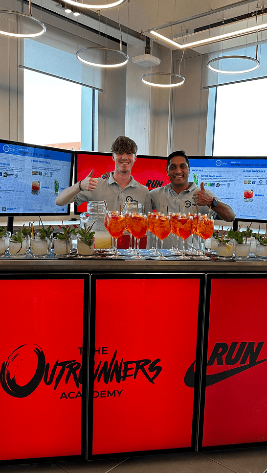 Two smiling bartenders giving thumbs up behind a glowing digital mobile bar displaying Nike and Outrunners Academy branding with fresh cocktails lined up on the counter
