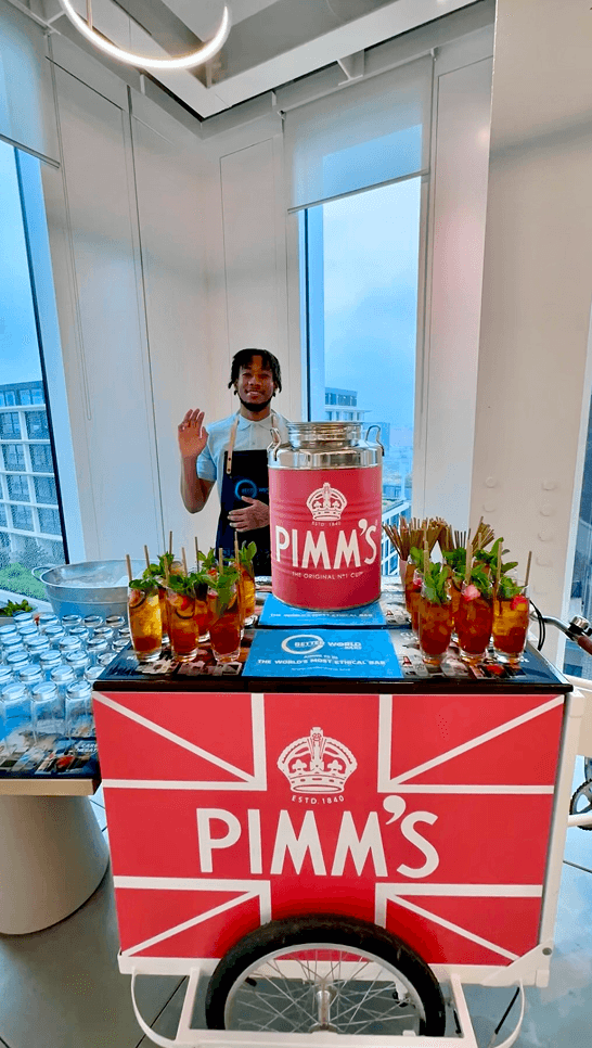 A smiling bartender wearing a denim apron waving from behind a branded Pimms tricycle loaded with fresh cocktails