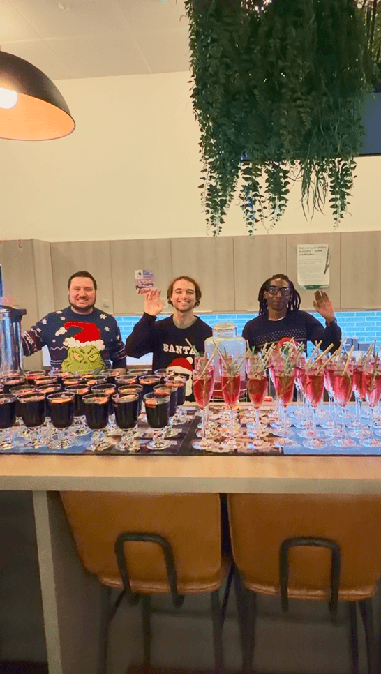 Three event bartenders wearing Christmas jumpers waving behind a long wooden table filled with rows of festive cocktails and mulled wine