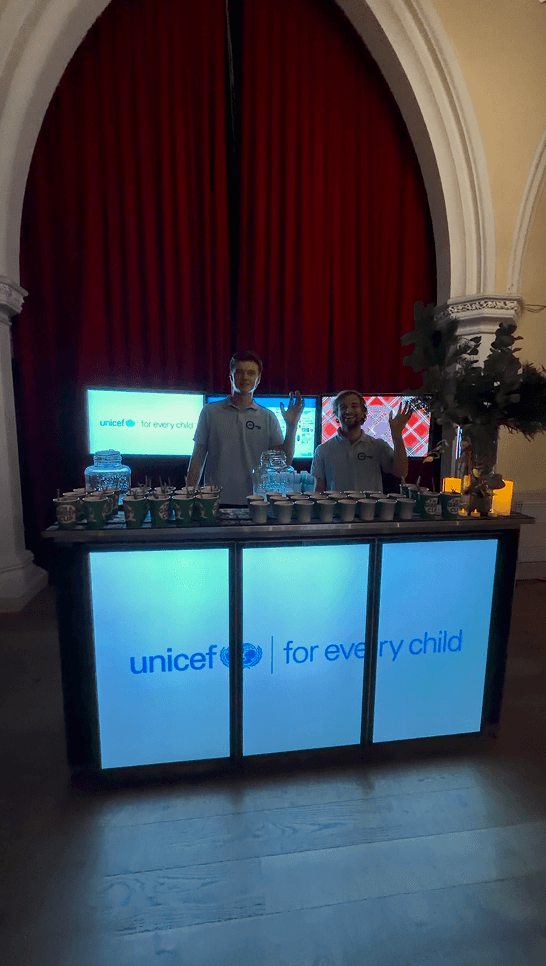Two smiling bartenders waving behind a glowing digital mobile event bar displaying the unicef logo at a charity fundraiser in London
