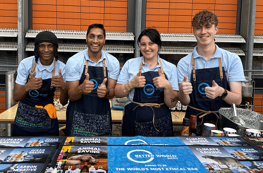 Four smiling event bartenders wearing denim aprons giving thumbs up behind a premium mobile bar