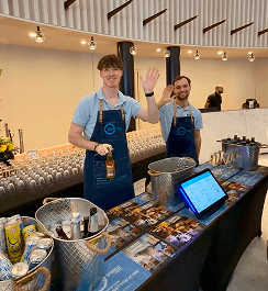 Two professional team members waving from behind an affordable pop up office bar setup serving premium drinks for a corporate team building event