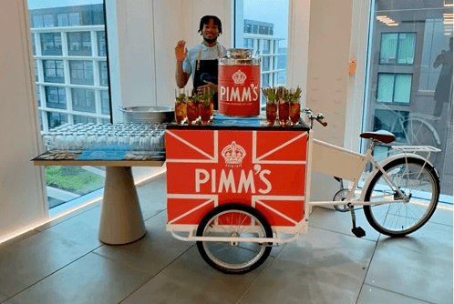 A professional bartender smiling and waving behind a branded Pimms tricycle serving summer cocktails at a corporate event in a modern office space.