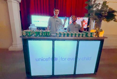 Two professional bartenders waving behind a digital event bar displaying the UNICEF logo. The bar is stocked with eco friendly cups and prepared for a charity fundraiser event.