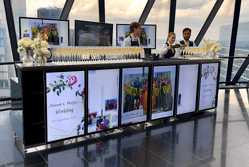 Two elegant team members pouring champagne behind a sophisticated digital wedding bar inside a luxury London venue with panoramic city views.