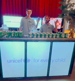 Two professional bartenders smiling behind a digital event bar displaying charity branding with eco friendly cups ready for service.