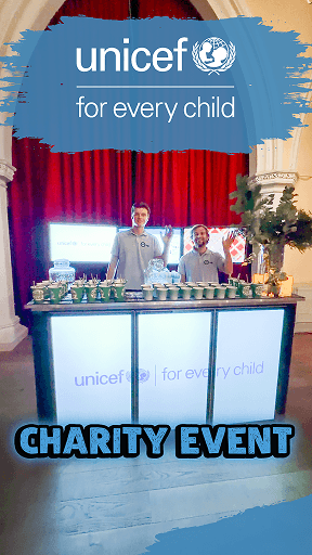 Two event staff waving from behind a custom branded digital mobile bar at a UNICEF charity event, serving drinks in eco-friendly disposable cups.