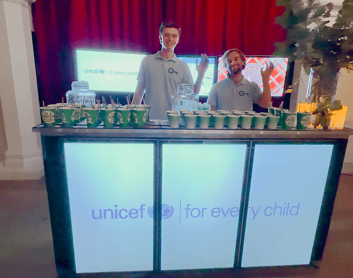 Two smiling team members standing behind a glowing digital mobile event bar displaying the UNICEF logo across its front panels representing our premium hospitality and mission driven corporate partnerships