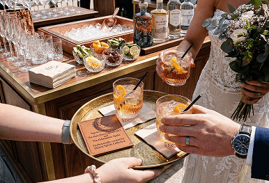A bride and a wedding guest taking fresh cocktails from a golden tray in front of a fully stocked outdoor wooden bar setup representing our seamless wedding bar logistics