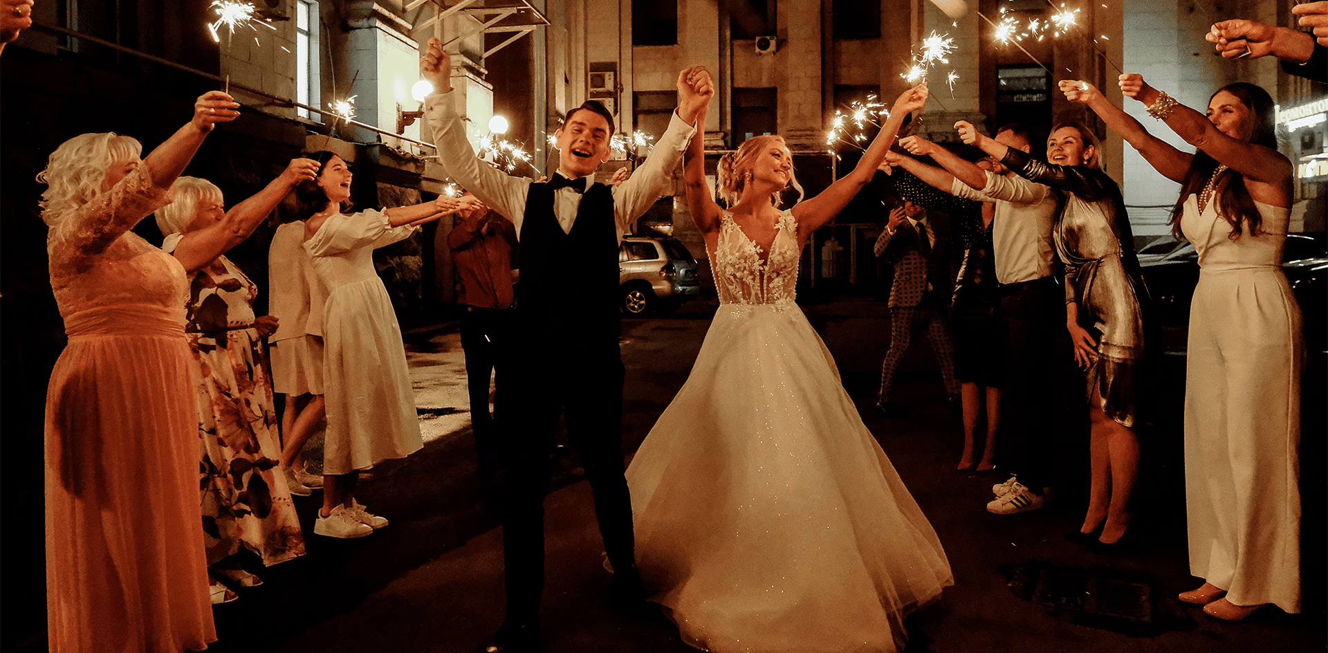 A joyful newly married couple walking through a sparkler exit and surrounded by cheering guests holding sparklers and light sticks at night representing the worlds most ethical wedding bar service for sustainable wedding bar hire THE WEDDING EXPERTS BEHIND YOUR SUSTAINABLY EVER AFTER