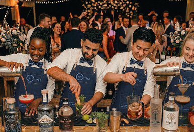 Four smiling professional bartenders mixing and pouring cocktails behind a rustic wooden bar at a lively wedding reception representing our mission driven mixology team