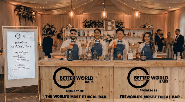 Four smiling bartenders raising colourful cocktails behind a rustic reclaimed wood eco wedding bar representing our sustainable bar hire service for marquee barn and woodland ceremonies