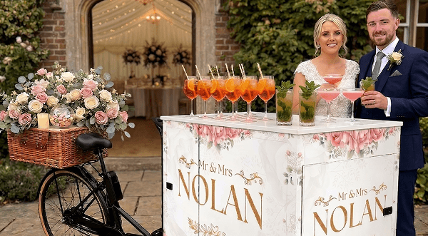 A newly married couple smiling and holding cocktails next to a beautifully decorated boutique wedding drinks tricycle cart on an outdoor patio representing our charming welcome drinks service