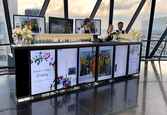 Three bartenders pouring champagne and preparing drinks behind a premium digital mobile wedding bar displaying personalised photos inside The Gherkin representing our five star service and sustainable wedding experts