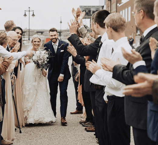 A happy newly married couple walking down an outdoor path while their smiling guests clap and cheer representing a five star review for our sustainable wedding eco bar from John and Michelle