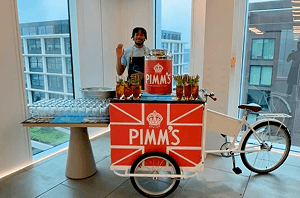 A smiling bartender waving from behind a Pimms branded tricycle mobile bar serving fresh cocktails in a modern corporate office