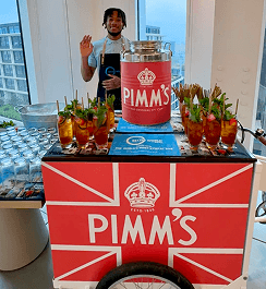 A smiling bartender waving from behind a Pimms branded drinks tricycle decorated with a British flag design and fresh fruit cocktails
