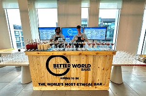 Two smiling bartenders standing behind a rustic wooden mobile bar covered in freshly made cocktails