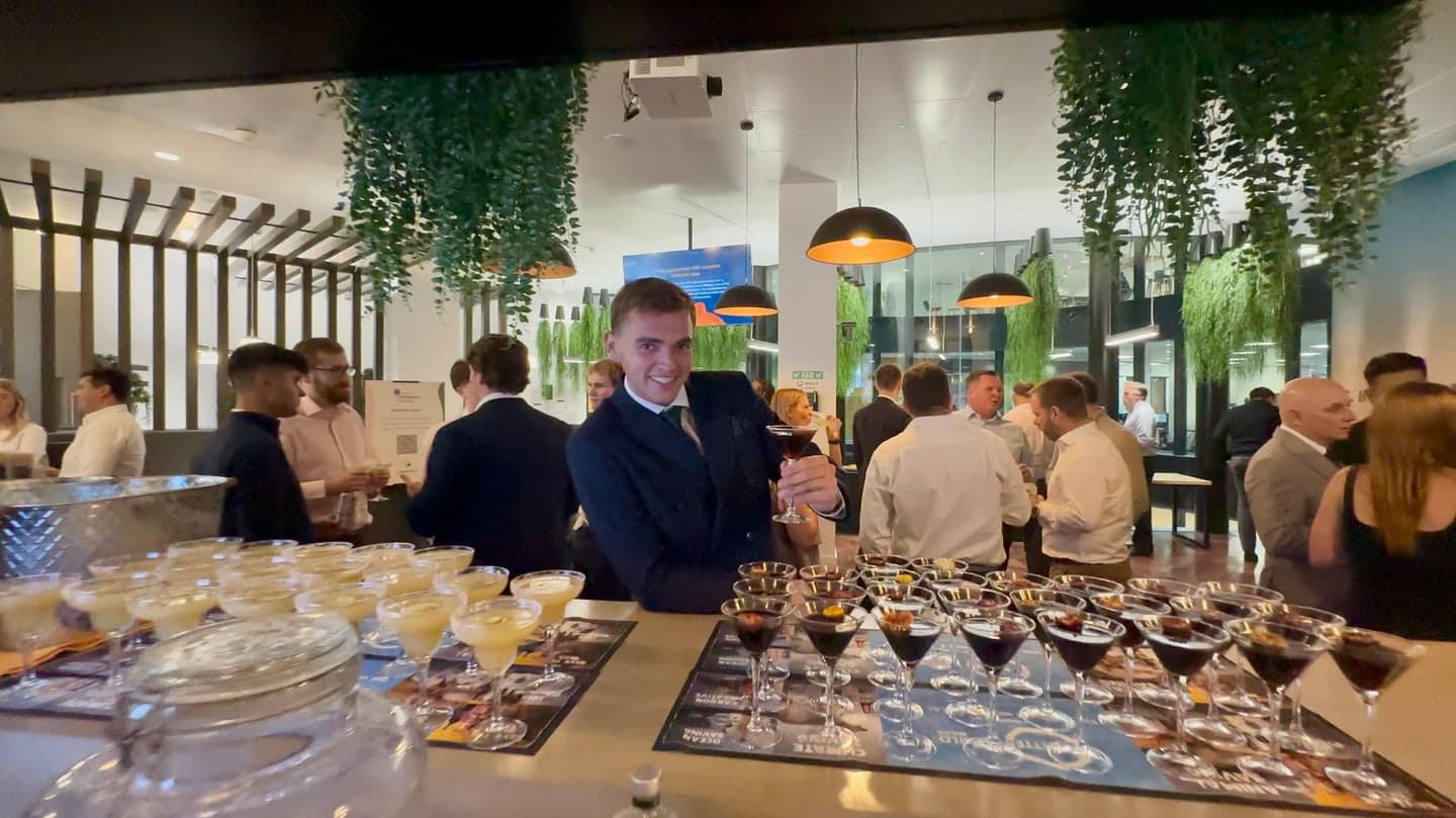 A smiling sustainability manager in a suit holding a cocktail beside colleagues at a corporate event bar