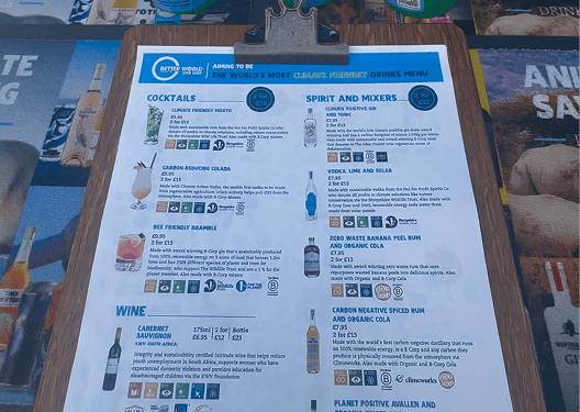 A wooden clipboard displaying a climate friendly drinks menu sitting in front of three fresh cocktails in green eco cups