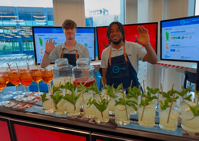 Two smiling bartenders wearing denim aprons waving from behind a premium mobile event bar lined with fresh cocktails