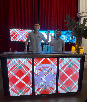 Two smiling bartenders waving from behind a glowing digital mobile bar displaying a red tartan pattern and the St Andrews Charity Ball logo